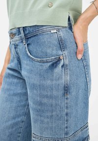 Helle blaue Denim-Jeans mit einem ausgewaschenen Finish, frontalen Taschen und sichtbaren Nähten. Der Bund hat einen Metallknopfverschluss.