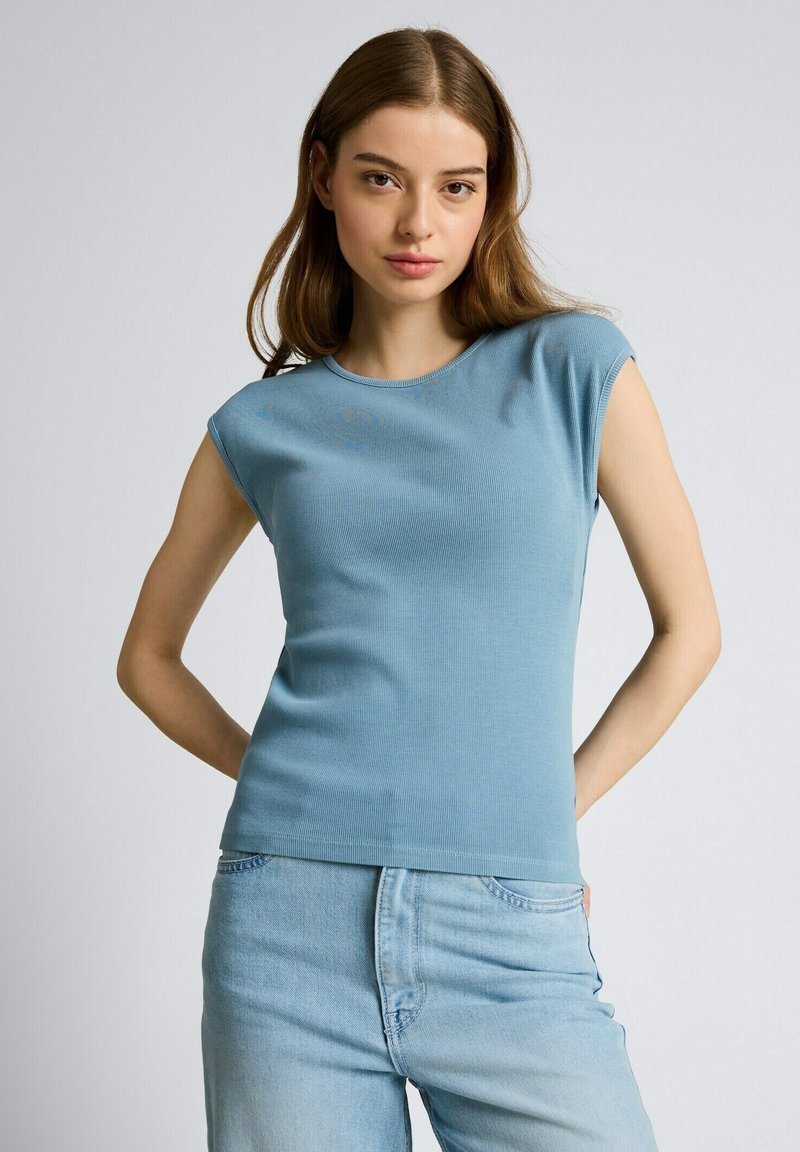 Junge Frau mit langen braunen Haaren, die ein ärmelloses hellblaues Oberteil und hellblaue Jeans trägt, steht vor einem schlichten Hintergrund.