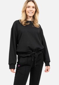 Felpa nera con vestibilità rilassata, polsini a costine e orlo con cordino; abbinata a pantaloni jogger coordinati con tasche laterali e dettagli sottili.
