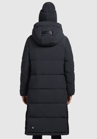 Abrigo acolchado negro largo con capucha, que presenta un diseño acolchado horizontal, aberturas laterales y una placa de logo cerca del dobladillo. Combinado con un gorro de lana.