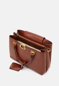 Sac à main en cuir synthétique marron avec un design structuré, une quincaillerie dorée, deux poignées supérieures et une petite étiquette attachée avec un détail de logo.