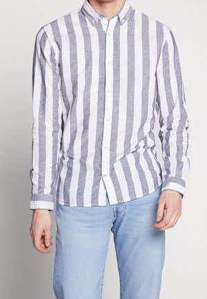 Homme portant une chemise boutonnée à rayures verticales blanches et grises avec un jean bleu clair sur un fond uni.