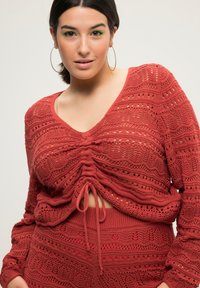 Rostfarbener Strickoberteil mit V-Ausschnitt, gerafftem Vorderteil und Kordelzugdetails. Mit offenen Mustern und langen Ärmeln.