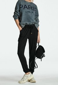 Sweat-shirt gris avec le texte "PARIS L'amour", pantalon noir fuselé avec poches, baskets beiges et un petit sac à dos noir texturé.