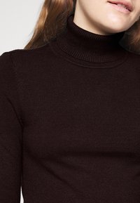 Maglione a collo alto di colore marrone scuro, realizzato in tessuto a costine, con vestibilità slim e colletto alto, dalla texture liscia e morbida.