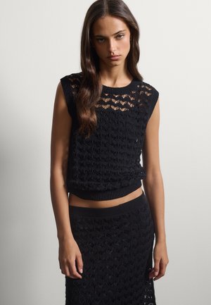 Giovane donna con top nero senza maniche in maglia e gonna coordinata in maglia, in piedi contro una parete bianca semplice.