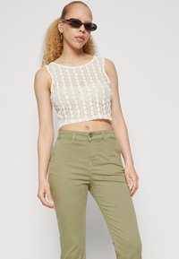 Krystall lace cropped topp i elfenben med et mønster av sirkulære blomster; kombinert med tettsittende olivengrønne bukser. Tilbehøret inkluderer mørke solbriller.