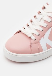 Rosa lädersneaker med vit gummisula, med vita snörningar och en logotypaccent. Designen inkluderar ett framträdande vitt sidostrek.