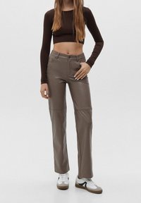 Braunes langärmeliges Crop-Top kombiniert mit beigen, geraden Kunstlederhosen. Das Model trägt weiße und graue Sneaker und steht vor einem schlichten Hintergrund.
