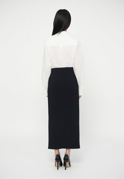 Γυναίκα με μακριά μαύρα μαλλιά, φορώντας λευκό πουκάμισο με μακριά μανίκια, χτυπημένο σε μια ψηλόμεση μαύρη midi φούστα και μαύρα ψηλοτάκουνα παπούτσια.