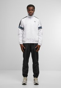Vit zip-jacka med marinblå ränder och svarta accenter, parat med svarta joggers och krämfärgade sneakers. Slät textur, atletisk passform.