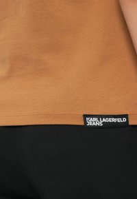Bruine shirtzoom met zwart "Karl Lagerfeld Jeans" label, gedragen over zwarte broeken, waarbij een deel van de zijkant en heup van een persoon zichtbaar is.
