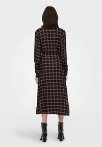 Frau in einem langärmligen, schwarz-roten karierten Midikleid mit einem Gürtel, kombiniert mit schwarzen Stiefeln bis zum Knöchel, die mit dem Rücken zum Betrachter steht.