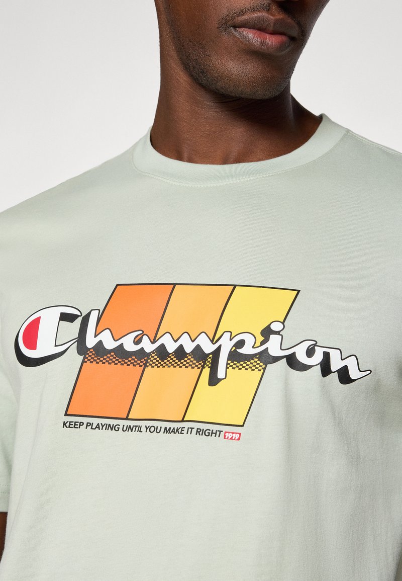 Jasno miętowa koszulka z grafiką w pomarańczowe i żółte pasy oraz napisem "Champion" i hasłem poniżej w kolorze czarnym.
