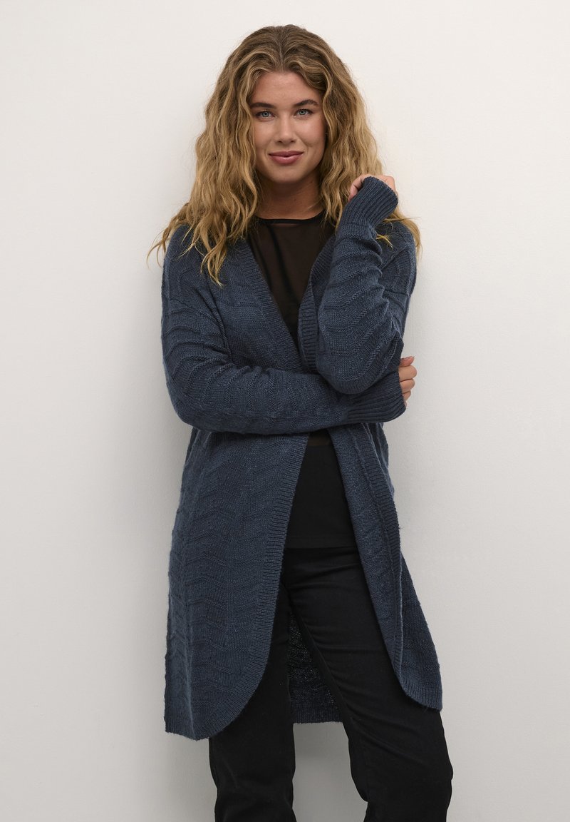 Lang navy cardigan laget av strukturert strikk, med V-hals-design, åpen front og ribbede ermer, båret over en svart skjorte og bukser.