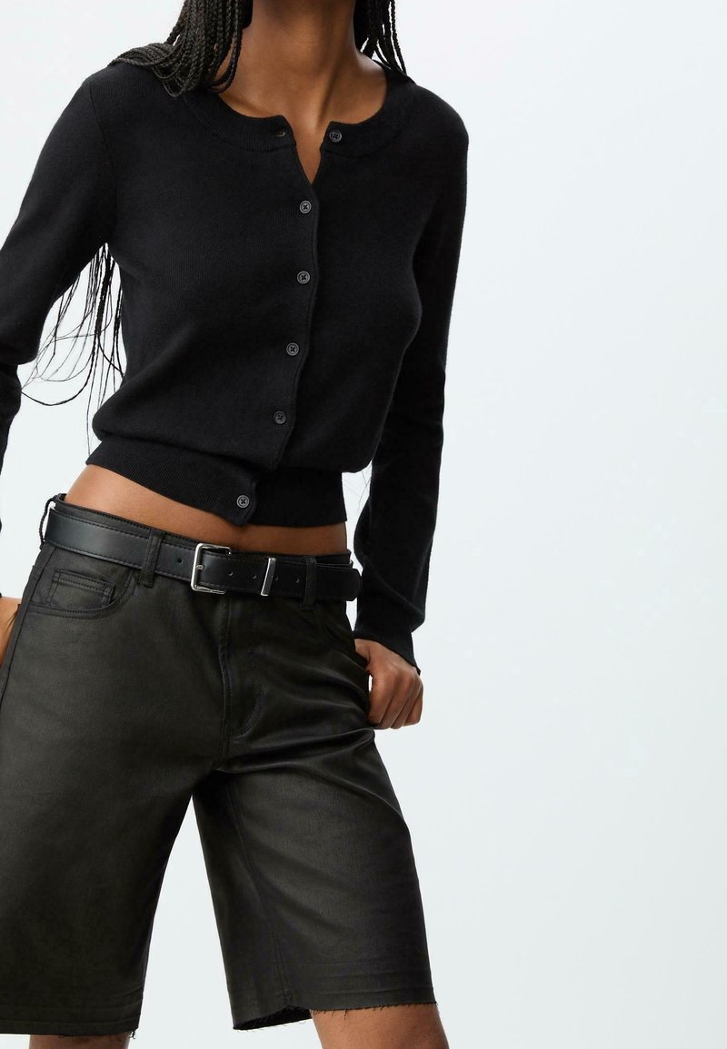 Femme portant un cardigan noir boutonné et un short en cuir noir taille haute avec une ceinture noire, posant devant un fond uni.
