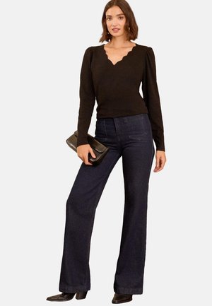 Frau trägt ein schwarzes, gewelltes V-Ausschnitt-Oberteil, dunkle weite Jeans, schwarze spitze Stiefel, hält eine schwarze Clutch.