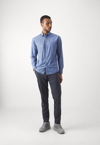 Camisa azul de botones con un patrón sutil, combinada con pantalones gris oscuro y zapatillas deportivas gris claro. De pie con las manos en los bolsillos.