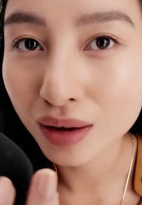 Gros plan d'une personne au teint lisse, cheveux foncés et couleur de lèvres neutre, tenant une éponge à maquillage noire avec un accent subtil sur les traits du visage.