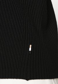 Tissu en maille côtelée noire avec une petite étiquette verticale présentant des rayures beige, blanche et noire près du bord inférieur central.