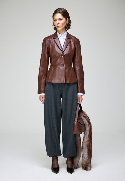 Femme portant une veste en cuir marron, pantalon large gris, chaussures pointues noires, tenant un sac à main marron avec un long accessoire en fausse fourrure.