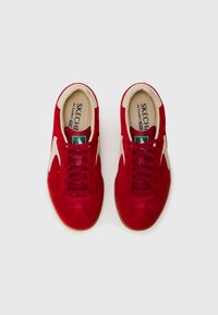 Røde semskede sneakers med beige detaljer, flade snørebånd og en tekstureret indersål. Har en gummisål og et distinkt paneldesign.