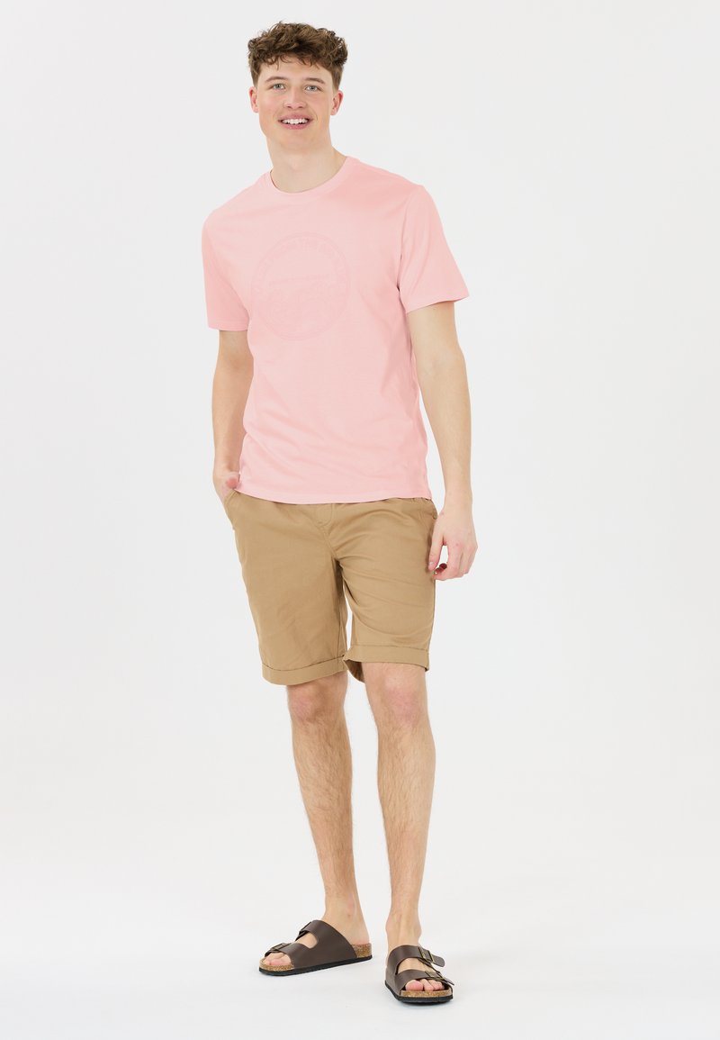 Vaaleanpunainen lyhythihainen t-paita, jossa on pyöreä graafinen kuvitus, yhdistettynä ruskeisiin shortsihousuihin ja ruskeisiin sandaaleihin, joissa on litteä hihna ja solkiyksityiskohtia.