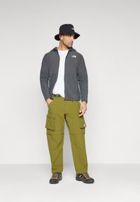 Gray zip-up jacket over a white t-shirt, olive green cargo pants with pockets, black bucket hat, and black hiking shoes with orange accents.