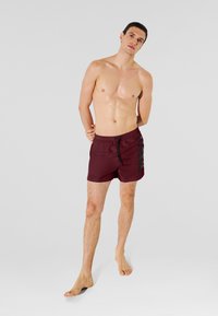 Bañador corto marrón granate con cordón negro y logo lateral. La tela es suave y ligera. El modelo está descalzo frente a un fondo gris.