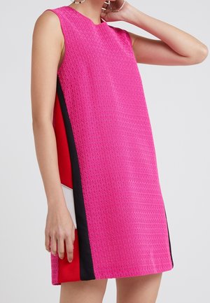 Robe rose sans manches avec un motif texturé, comportant des panneaux verticaux noirs, rouges et blancs sur les côtés, portée par une personne debout.