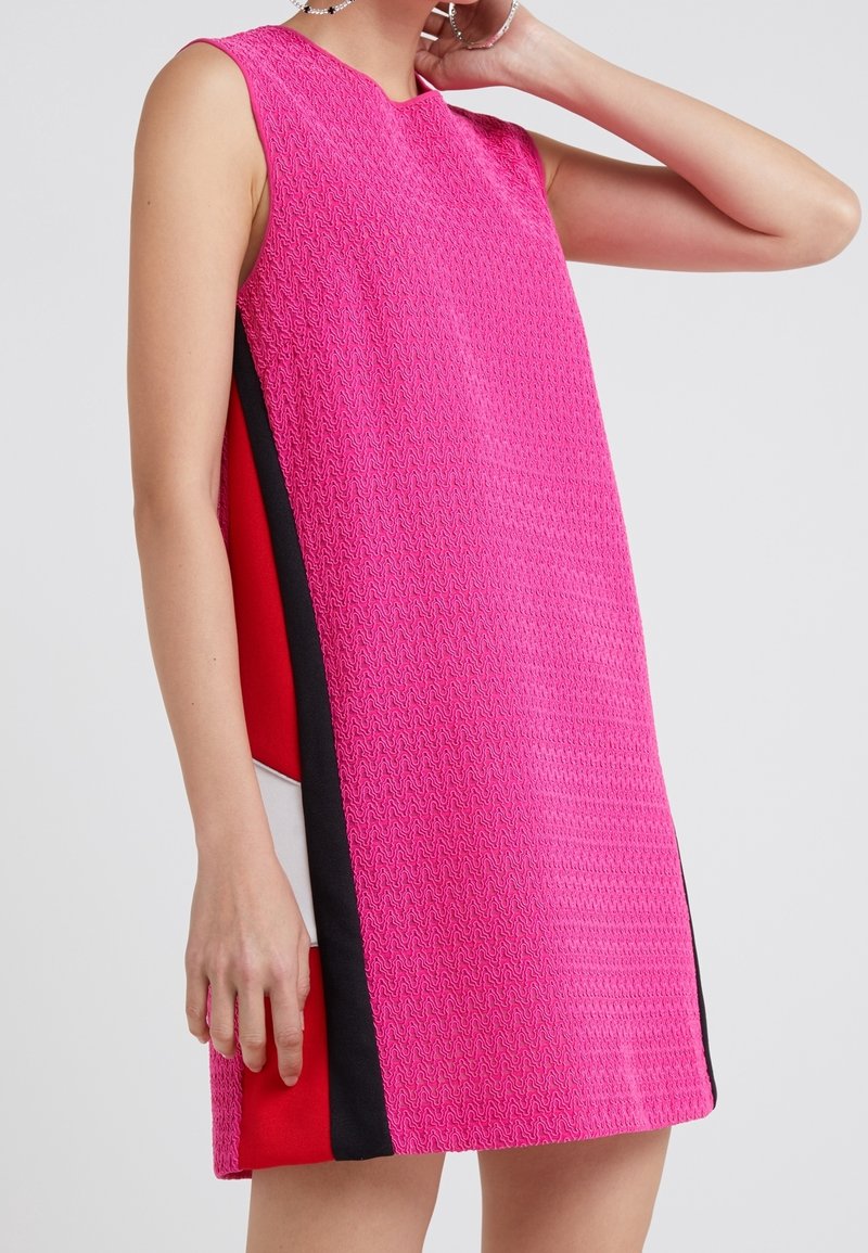 Robe rose sans manches avec un motif texturé, comportant des panneaux verticaux noirs, rouges et blancs sur les côtés, portée par une personne debout.