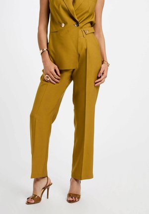 Completo giallo senape su misura con blazer doppiopetto, pantaloni larghi e dettagli in oro. Sandali alti con cinturini marroni.