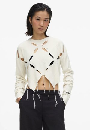 Femme aux cheveux courts et foncés porte un pull blanc à manches longues avec des découpes ovales et des bords effilochés, associé à un pantalon noir.