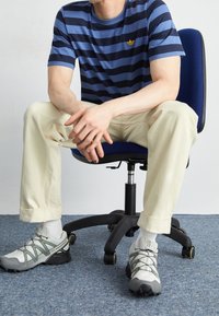 Person i blå och svartrandig skjorta och krämfärgade byxor som sitter på en blå kontorsstol med händerna knäppta, iförd vita och grå sneakers.