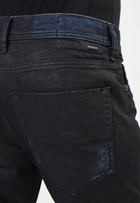 Detailní záběr osoby v černých denimových džínách s viditelnou zadní kapsou a malou kovovou etiketou značky Diesel na pase.