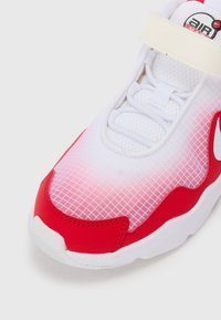 Zapatilla blanca con acentos rojos, que presenta una parte superior de malla, un patrón en cuadrícula y una correa sobre los cordones. Suela de goma y diseño de punta redondeada.