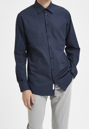 Man wearing a navy blue button-up shirt with small white geometric patterns and light gray pants, standing with one hand in pocket.