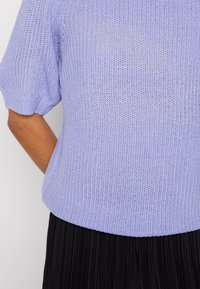 Person in einem hellpurpurnen, gestrickten Kurzarmpullover und einem schwarzen, gefalteten Rock, mit entspanntem Arm an der Seite vor einem weißen Hintergrund.