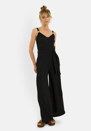 Mujer con el pelo rubio recogido en un moño que lleva un mono negro sin mangas con pantalones anchos y sandalias de plataforma marrones, posando frente a un fondo blanco liso.