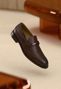 Pruun nahast loafers sileda viimistlusega, millel on metallist rihma aksent ja ümar varvas. Must kummist tald minimaalse mustriga.