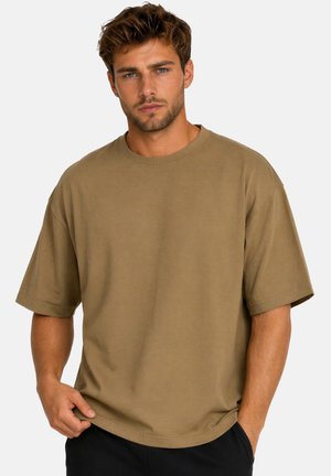Mężczyzna z krótkimi brązowymi włosami, ubrany w luźny, jednolity brązowy t-shirt i ciemne spodnie, stoi na jasnym tle z neutralnym wyrazem twarzy.