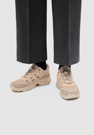 Baskets beiges à lacets avec semelles épaisses portées avec des chaussettes noires et un pantalon court gris foncé sur fond clair.