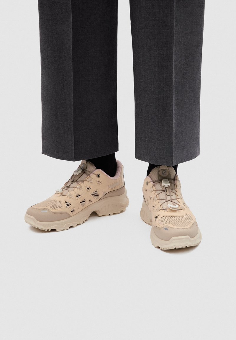 Baskets beiges à lacets avec semelles épaisses portées avec des chaussettes noires et un pantalon court gris foncé sur fond clair.