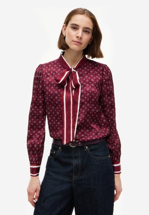 Femme portant un chemisier bordeaux à motifs avec un grand col à nœud papillon et un jean taille haute bleu foncé, debout devant un fond blanc uni.