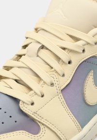 Közelkép egy bézs sportcipőről, amelynek oldalán és fűzőin világoskék átmenet található, bemutatva az ikonikus Nike logót.