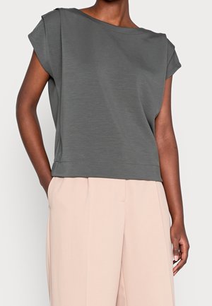 Femme portant un haut ample gris sans manches avec des épaules rembourrées et un pantalon beige clair taille haute, les mains partiellement dans les poches.