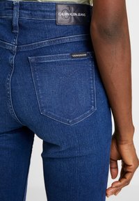 Mörkblå denimjeans med hög midja, som har en läderlogotyp och sömnadsdetaljer på bakfickan. Slät textur med lätt stretch.