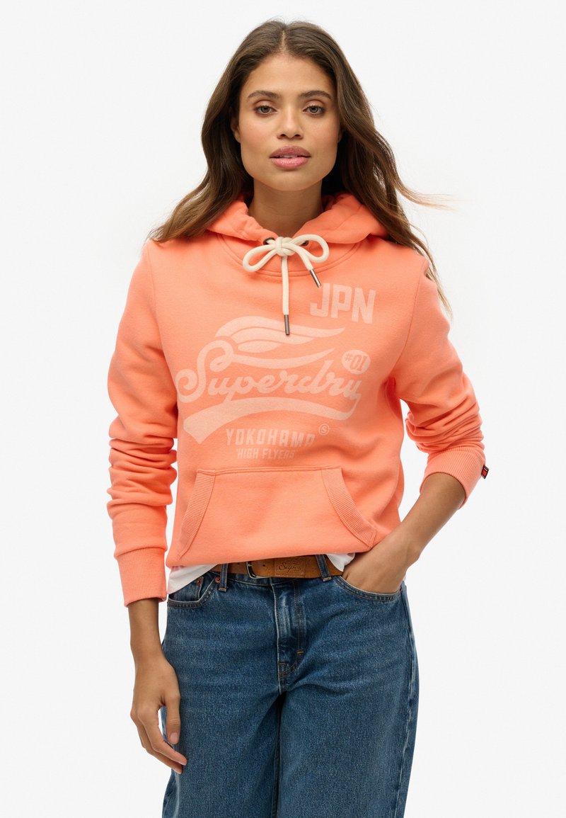 Sweat à capuche corail en tissu doux, avec une poche avant, un cordon de serrage noué et un imprimé graphique avec texte blanc. Porté avec un jean bleu.
