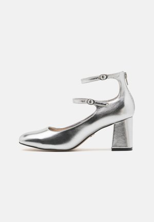 Chaussure à talon haut en métal argenté avec des brides de cheville, bout rond et talon bloc. Texture lisse avec une finition brillante et un détail à deux brides.