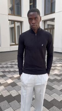 Black zippered sweater with long sleeves, featuring a small white logo. Paired with white, pleated trousers, standing on patterned pavement.